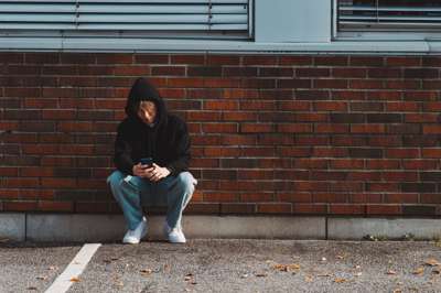 En tenåringsgutt  med olabukse og hettegenser sitter på huk med ryggen mot en rød murstein-vegg. Han ser på mobiltelefonen sin.